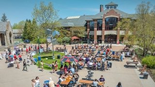 Students gather at outdoor tables for a spring event in front of Sullivan Student Center at Saint Lawrence University, with booths, banners, and trees in bloom nearby.