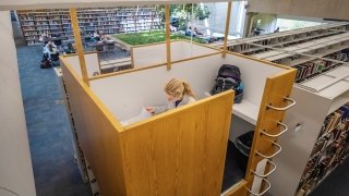 A student studies in a private wooden study pod elevated above library bookshelves at Saint Lawrence University, with open tables and bookshelves visible in the background.