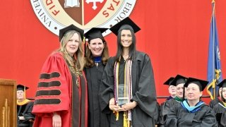 President Kate Morris, Jennifer Curley Reichert and a student stand together, wearing regalia, on the Commencement stage.