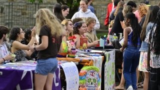 Students gather around tables at an outdoor student club fair at Saint Lawrence University, chatting and signing up for activities. Tables are decorated with colorful signs, posters, and props, including a piñata-style skull and a &quot;Photography Club&quot; banner.