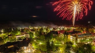 A nighttime aerial view of Saint Lawrence University’s campus lit with green trees and building lights, as red and gold fireworks burst in the sky above.
