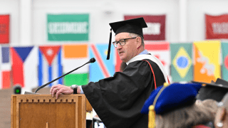 Michael Ranger, wearing graduation regalia, stands at a podium and address the crowd. Several countries flags are in the background.