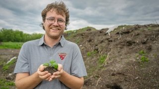 Sam Voter, wearing a grey Saint Lawrence University polo, holds a handful of dirt with a green sprout in the center, a large compositing pile is in the background.