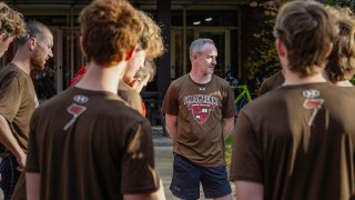 Paul Graham wearing a "Saint Lawrence Cross Country" shirt stands in the center of a group of team members during an outdoor meeting.