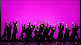Dance ensemble performs on stage against a bright pink backdrop.