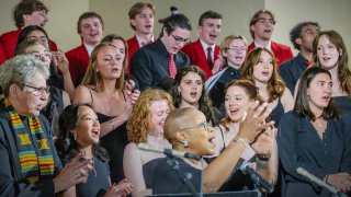A diverse group of students and a conductor perform together in a choir, singing enthusiastically.