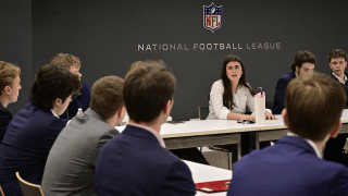 Students sit around a conference table in a meeting room with the National Football League logo on the wall. One person speaks while others listen, dressed in business attire. Water bottles, notebooks, and laptops are on the table.