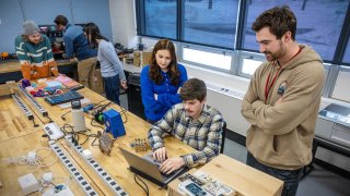 Students collaborate at a workbench in a Saint Lawrence University lab, using a laptop and electronic components. One student types while others stand nearby, observing and discussing the project. Tools, wires, and equipment are spread across the table, showing hands-on learning in progress.