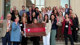 Group of adults stand on steps outside a building holding a Saint Lawrence University banner and red foam fingers. Many wear name badges and smile toward the camera. The group appears gathered for a campus or alumni event.