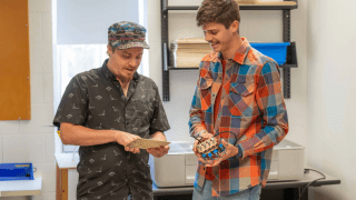 Two students stand in a lab space, examining electronic components and a small circuit board. One student holds a patterned board while the other holds a wired device, and both look down at the materials with focused expressions. Shelving and equipment are visible behind them, suggesting a hands-on learning environment at Saint Lawrence University.