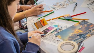 Two people sit at a table creating a collage with small printed photos, colored pencils, and tape. One person draws in a notebook while arranging images of landscapes and sunsets. Art supplies and cut paper pieces are spread across the workspace.