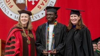 A male student holding an awards stands between two women in graduation regalia.
