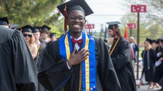 A student in a cap, gown, and blue stole smiles while preparing for his Commencement ceremony.