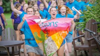 Students cheer while carrying a colorful handmade banner during an outdoor campus event.