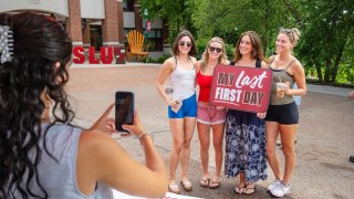 Four individuals pose outdoors holding a sign that reads "My Last First Day" while another person captures the moment with a smartphone.