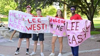 Students hold colorful handmade signs welcoming new residents to Lee North, Lee West, and Lee East on a tree-lined campus walkway.