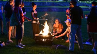 Students gather around a fire pit at dusk, roasting marshmallows together on a campus lawn.