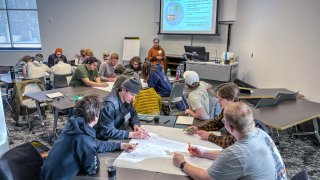 Students work together on a large group activity at classroom tables while a facilitator gives instructions at the front of the room.