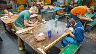Several people are working together in a workshop, carving and shaping wooden boards at large tables surrounded by wood shavings and tools.