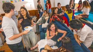 A group of young people socialize in a room with flags, eating snacks and chatting around couches and a pool table.