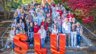 A group of Saint Lawrence alums and current students stand in front of the student center on a sunny fall day. There are large SLU letters in front of the group.