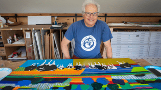 Woody Jackson, wearing a blue T-shirt stands behind a brightly colored painting featuring abstract shapes, bold stripes, and black-and-white cows in an art studio.