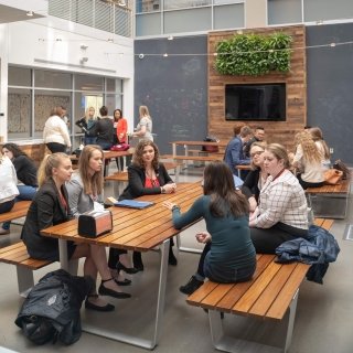 A group of students and alumni engaged in a networking conversation at a wooden table in a stylish office cafeteria.