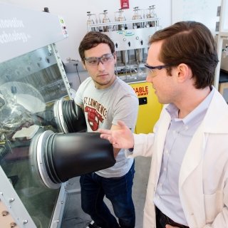 A student and professor, both wearing protective goggles, perform a chemistry experiment.