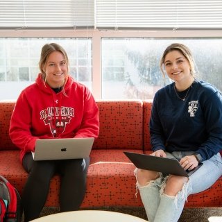 Two students sit on a red couch in a bright, airy study space with lots of windows. They both type on laptops.
