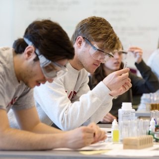 Three students sit at a bench in the science lab. They are all wearing safety goggles and prepping test tubes for an experiment.