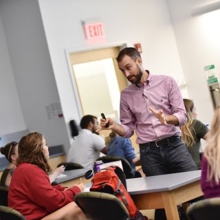 A teacher wearing a light red button-down shirt stands among his students while explaining a concept.