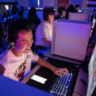A student, wearing a white Saint Lawrence e-sports jersey and headphones, smiles at a lit up computer screen and taps on a back-lit keyboard. Several students wear headphones and sit at computers in the background in a dimly lit room.