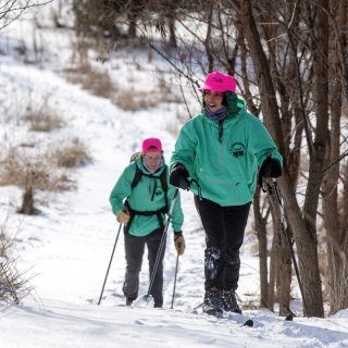 Two students, wearing sea foam green jackets and neon pink hats, cross country ski up a hill.