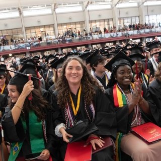 A group of Saint Lawrence students, wearing graduation regalia, sit in the front row at Commencement smiling as they flip their tassles.