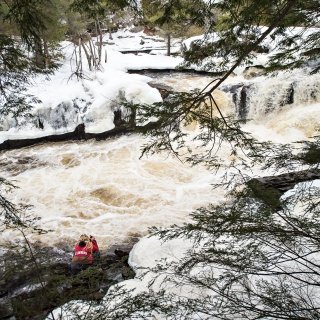 A student, wearing a scarlet Saint Lawrence rain jacket, crouches by a roaring river and takes photos.