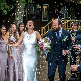 A bride, wearing a satin gown, holds a silver cup in her hand and laughs alongside her husband as they walk through the receiving line while flower petals fall from the sky. A brown cabin is in the background.