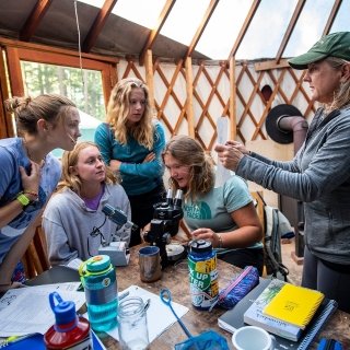 A professor holds up a piece of paper in explanation to a group of students in a yurt.