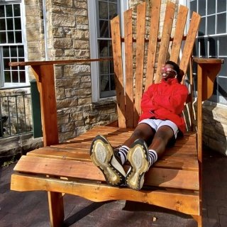 A Saint Lawrence student, wearing a scarlet zip up jack and solar sunglasses, leans back in a giant adirondack chair and smiles up at the son.