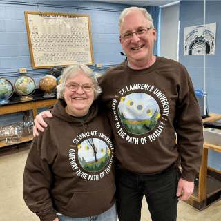 Aileen O'Donoghue and Jeffrey Miller wear brown Saint Lawrence University Gateway to the Path of Totality sweatshirts and stand in a physics lab.