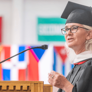 A person in a graduation cap and gown stands at a wooden podium, speaking into a microphone. The individual, who appears to be a faculty member or guest speaker, has white hair and wears glasses. In the background, colorful flags representing various countries or institutions are slightly blurred, creating a vibrant backdrop for the scene. The speaker's expression is focused and engaging, emphasizing the importance of the occasion.