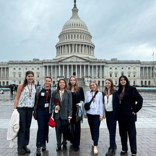 A group of seven people stands in front of the United States Capitol building on a cloudy day. They are all dressed in business casual attire and wearing conference lanyards around their necks. The ground is wet, indicating recent rain.