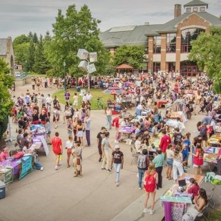 A large outdoor event at Saint Lawrence University, featuring many tables with colorful displays and students interacting. The scene is busy with people walking around, sitting at tables, and exploring various booths. A brick building and trees are visible in the background.