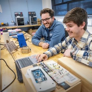 Two people sit at a workbench in a lab, smiling while working on a laptop connected to an electronic circuit project with wires and components.