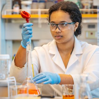 A Saint Lawrence University student, wearing a white lab coat, blue rubber gloves, and protective eye wear, measures liquid in a beaker in a chemistry lab. There are several measurement tools and beakers around them.