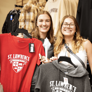 Two smiling people hold up Saint Lawrence University Saints shirts in a campus store, surrounded by racks of clothing.