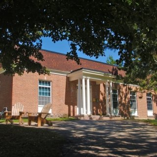 Gaines Hall at Saint Lawrence University is a red-brick building with white columns and large windows. The front lawn includes wooden chairs for outdoor seating under the shade of nearby trees. The building has a classic look and is surrounded by green space.