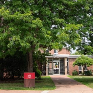 Lee Hall at Saint Lawrence University is a red-brick building surrounded by trees and green space. A path leads to a glass entryway, and outdoor seating is available with benches and Adirondack chairs. A red sign with white text marks the building’s name.
