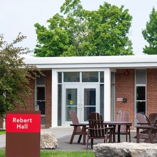 Exterior of Rebert Hall with a red-brick facade, glass entrance doors, a red sign, and outdoor seating with Adirondack chairs and tables.