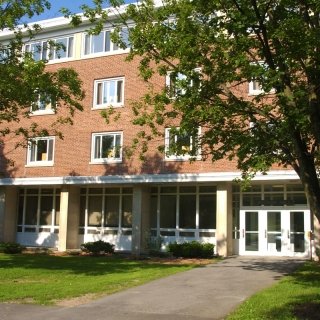 Exterior view of Whitman Hall, a red brick building with large white-trimmed windows and a main entrance framed by columns, partially shaded by green leafy trees on a sunny day.