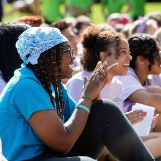 A group of students sitting outside on a sunny day, smiling and clapping during an event at Saint Lawrence University.